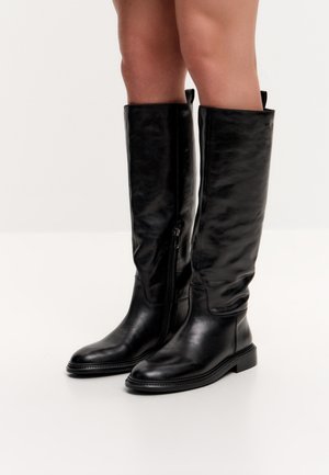 Schwarze kniehohe Lederstiefel mit seitlichen Reißverschlüssen, getragen von einer Person, die auf einer weißen Oberfläche steht.