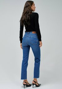 Jeans de tiro alto azules con pierna recta y dobladillo deshilachado, combinados con una camiseta negra de manga larga. Parche de marca de cuero marrón distintivo en la parte trasera.