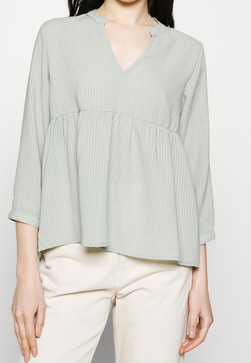 Femme portant un chemisier texturé vert clair avec un col en V et des manches trois-quarts, associé à un pantalon beige clair, debout devant un fond blanc.