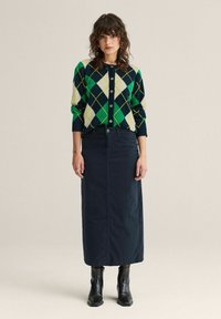 Neulecardigan vihreällä, laivastonsinisellä ja kerma-valkoisella argyle-kuviolla, yhdistettynä pitkään laivastonsiniseen hameeseen ja mustiin nilkkureihin. Nappilista edessä.