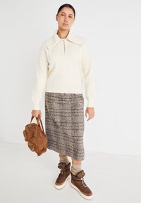 Elfenbenfarget strikkegenser med stor krage og glidelås, kombinert med et brunt rutete midiskjørt. Brun veske og chunky brune sneakers fullfører antrekket.