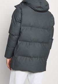 Chaqueta acolchada de color gris oscuro con capucha, que presenta secciones acolchadas para mayor aislamiento. Textura suave, mangas largas y silueta ajustada.