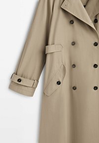 Trench-coat beige double boutonnage avec boutons noirs, sangle de manche réglable et rabat de poche latérale boutonné.