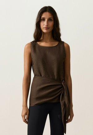 Donna con lunghi capelli scuri che indossa una canotta marrone senza maniche con fiocco laterale e pantaloni neri, in piedi davanti a uno sfondo chiaro e semplice.