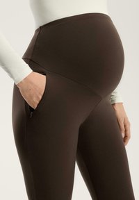 Hnědé těhotenské legíny s vysokým pasem, vyrobené z elastického materiálu, s kapsou na zip na boční straně a hladkým povrchem.