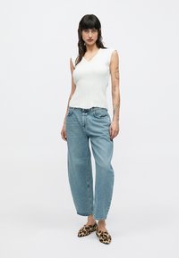 Γυναίκα που στέκεται φορώντας λευκή αμάνικη μπλούζα, φαρδιά μπλε τζιν και μυτερά παπούτσια με εκτύπωση λεοπάρ, σε απλό φωτεινό φόντο.