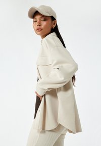 Beige oversized jakke med lange ærmer og en klaplomme. Båret over en mørk bluse og parret med lyse ribbede bukser og en matchende kasket.