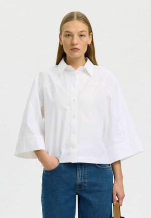 Mladá žena s dlhými rovnými vlasmi, oblečená v voľnej bielej košeli na gombíky a modrých džínsoch, stojaca s jednou rukou v vrecku.