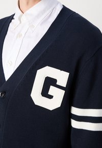 Marinblå stickad cardigan med en vit "G"-patch och två vita ränder på ärmen. Har en V-ringning och knappstängning, i kombination med en vit skjorta.