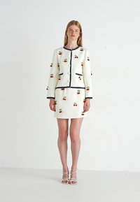 Mujer vestida con un traje de falda blanco con pequeños patrones de cereza, ribete azul marino y sandalias blancas con tiras, de pie frente a un fondo blanco plano.