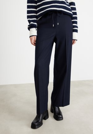 Persoon draagt marineblauwe wijde broek met trekkoord in de taille, zwarte chunky haklaarzen en een marineblauw-wit gestreepte trui.