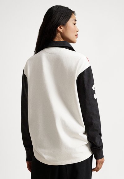 Chemise à manches longues en coton blanc et noir. Elle présente un col avec collet, des manches contrastantes, et un grand graphique numérique sur le bras.