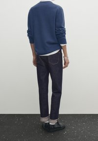 Pull en maille bleu, sous-vêtement blanc, jean en denim foncé avec revers roulés, et chaussures noires. Coutures visibles sur le pantalon et texture côtelée des manches.