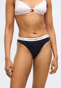 Sieviete, tērpusies baltā krūšturī un melnā Tommy Hilfiger apakšveļā ar zīmola elastīgu jostu, stāv pret vienkāršu gaišu fona sienu.