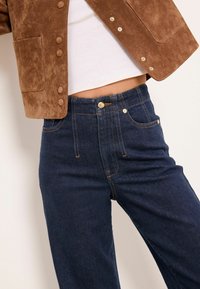 Barna szarvasbőr dzseki patent gombokkal, sötétkék, magas derekú farmerrel párosítva, amely aranyszínű fém részletekkel és kontrasztos varrással rendelkezik.