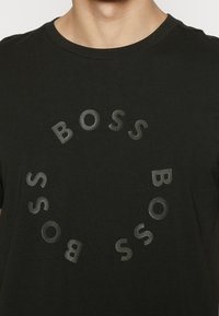 Delvis vy av en person som bär en svart t-shirt med perforerade bokstäver som stavar "BOSS" i ett cirkulärt mönster på bröstet.