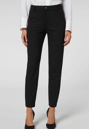 Vrouw draagt nauwsluitende zwarte broek, wit overhemd met knopen en zwarte hoge hakken, staand tegen een lichtgrijze achtergrond.