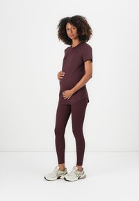 Gravid kvinne som står og holder på magen, iført en matchende mørk lilla antrekk og hvite Nike-sneakers mot en hvit bakgrunn.