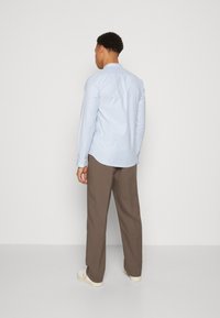 Camicia a maniche lunghe blu chiaro con orlo curvo e polsini con bottone, abbinata a pantaloni marroni e scarpe bianche, vista da dietro.