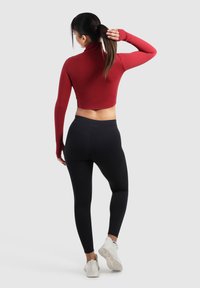 Rode cropped top met lange mouwen en hoge kraag, gecombineerd met zwarte leggings, gezien vanuit de rug. Witte sportschoenen maken de outfit compleet.