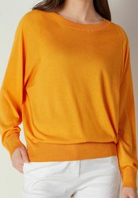 Femme portant un pull à manches longues d'un orange vif, légèrement rentré dans un pantalon blanc, avec une main dans la poche, sur un fond neutre.