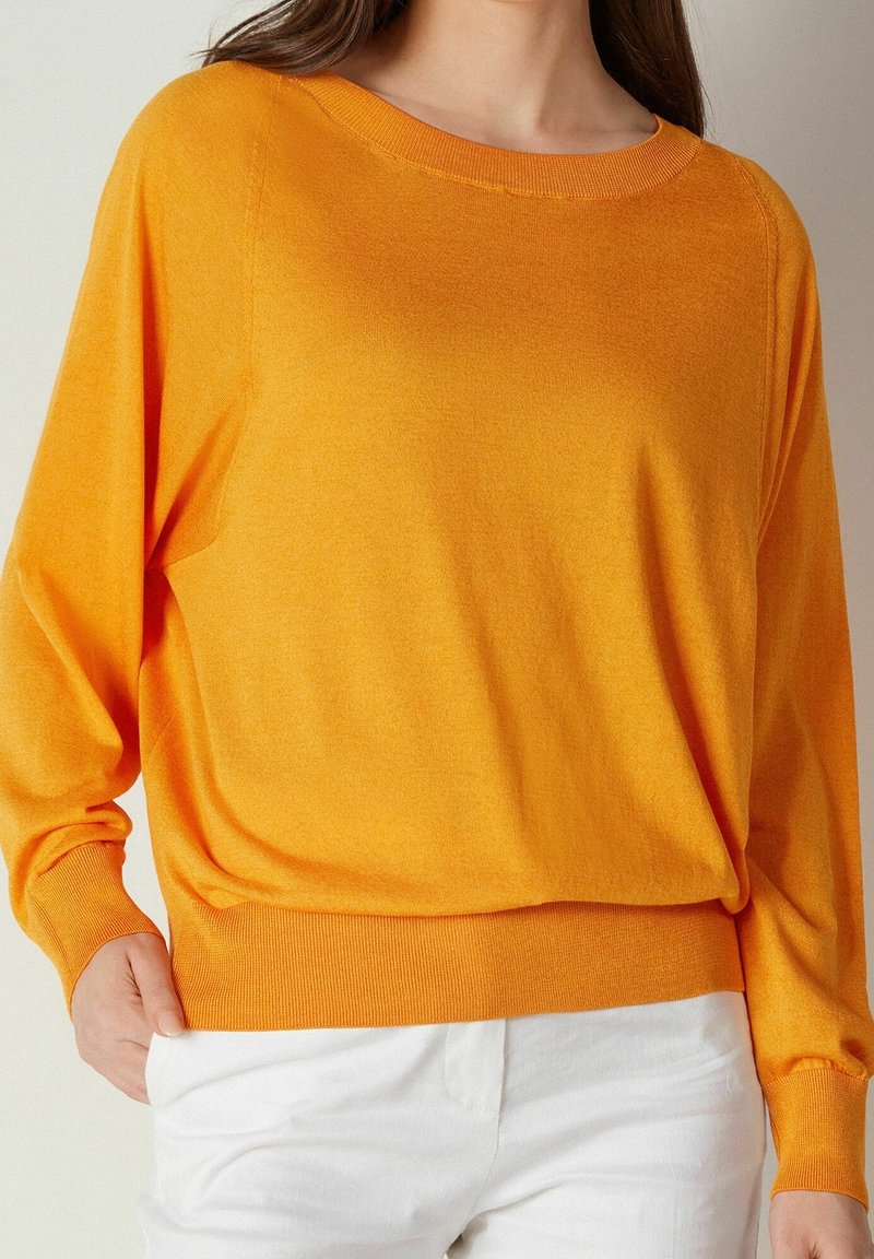 Femme portant un pull à manches longues d'un orange vif, légèrement rentré dans un pantalon blanc, avec une main dans la poche, sur un fond neutre.