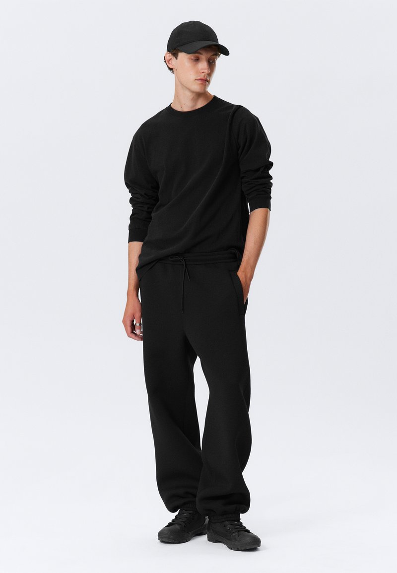 Chemise noire à manches longues et pantalon de survêtement, tous deux en tissu doux et texturé. La tenue est complétée par une casquette de baseball noire et des baskets noires.