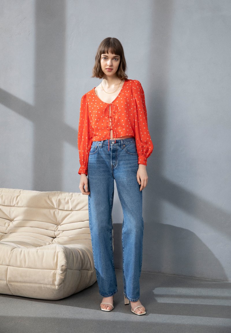 Blusa vermelha com bolinhas brancas, com mangas balão e um laço na frente, combinada com jeans retos de cor azul claro e sandálias de salto alto bege com tiras.