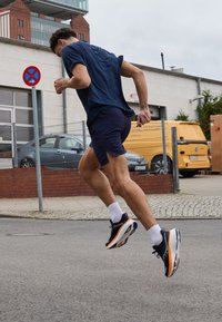 Idrettssko med svart overdel, orange detaljer og hvite såler; brukt av en person i en marineblå skjorte og shorts mens de løper utendørs.