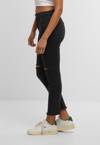 Černé džíny z denimu s roztrženými koleny, vysokým pasem a surovým lemem. Doplňují je bílé tenisky se zelenými akcenty a textovým detailem.