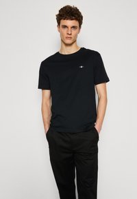 T-shirt en coton noir à manches courtes, col rond, avec un branding subtil sur la poitrine, associé à un pantalon noir. Design simple et coupe classique.