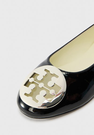 Zwarte lakleren ballerina met een ronde neus en een groot zilveren logo op de neuskap. De binnenkant is lichtbeige van kleur.