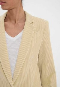 Blazer en velours côtelé beige avec des côtes verticales, col structuré et poche poitrine unique, associé à un t-shirt blanc à col en V en dessous.