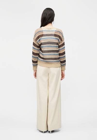 Γυναίκα που φαίνεται από πίσω, φοράει πολύχρωμο ριγωτό πλεκτό πουλόβερ και φαρδιά κρεμ παντελόνια σε απλό φόντο.