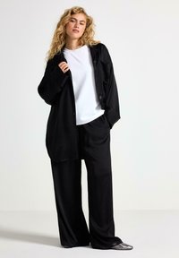 Camicia nera oversize con bottoni, t-shirt bianca a girocollo sotto, e pantaloni neri larghi. Modello che indossa scarpe argento metallico.