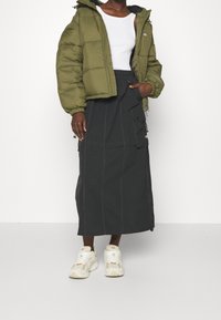 Giacca a vento verde oliva, canotta bianca, gonna midi nera con tasche e sneakers cremosi con dettagli marrone chiaro.