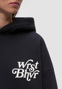 Granatowy hoodie z dużym białym logo "Wrst Bhvr" na przodzie. Charakteryzuje się szerokim, miękkim kapturem oraz teksturowanym wykończeniem materiału.