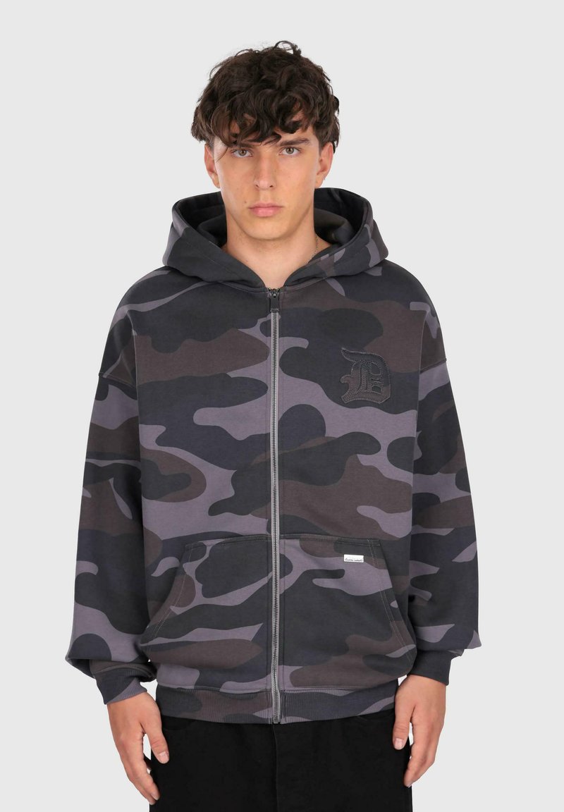 Ung mand iført en lynlåsbetjent mørkegrå og sort camouflage hoodie med hætte, stående foran en ensartet lys baggrund.