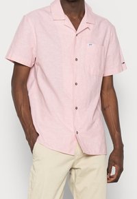 Uomo che indossa una camicia a maniche corte di colore rosa chiaro con taschino e pantaloni beige, in piedi con una mano in tasca.
