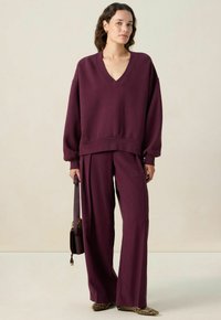 Vrouw in een losse bordeauxrode V-halstrui en brede broek, met een gestreepte handtas, staand tegen een effen lichte achtergrond.