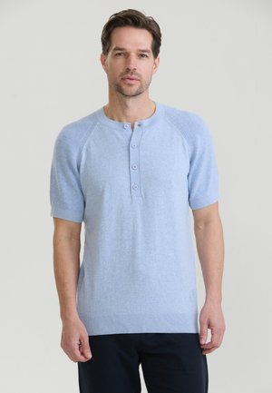 Mężczyzna ubrany w jasnoniebieską, krótki rękaw, zapinaną na guziki koszulę i czarne spodnie, stojący na gładkim jasnym tle.