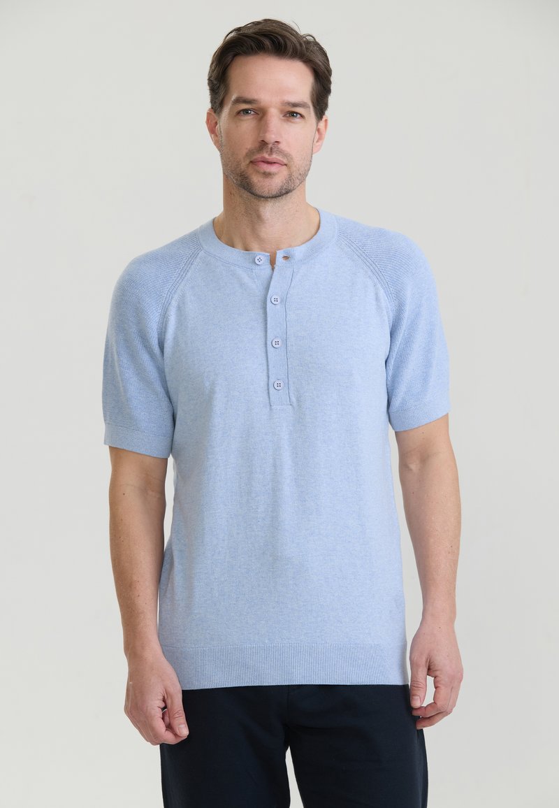 Homme portant une chemise bleu clair à manches courtes boutonnée et un pantalon noir, debout devant un fond clair uni.