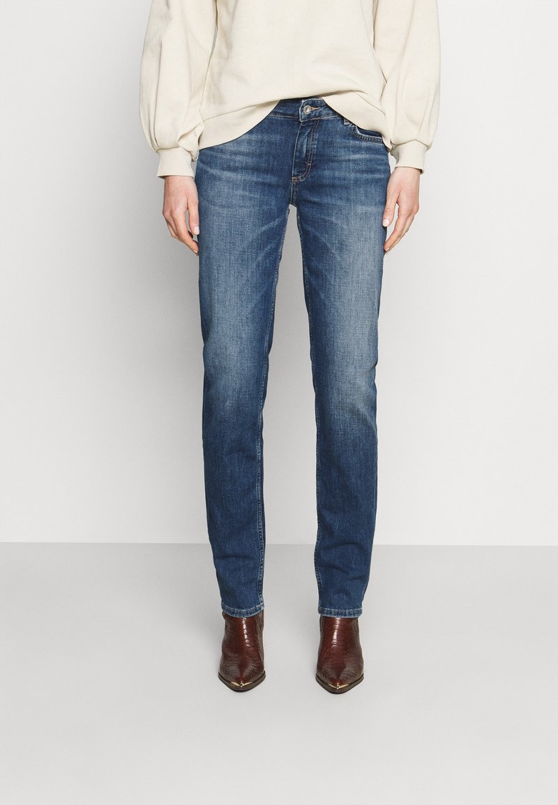 Femme portant un jean en denim bleu, des bottes marron à bout pointu, et un chemisier crème à manches longues, debout devant un fond clair uni.