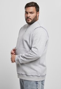 Grå hettegenser med rund hals, ribbestrikkede ermer og nederkant. Har kontrasterende sømmer langs skuldrene. Brukt med lyseblå jeans.