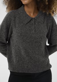 Pilkas megztinis su apykakle, keturiais juodais mygtukais ir švelnia tekstūra. Siluetas laisvas, su nerti krašto detalėmis.
