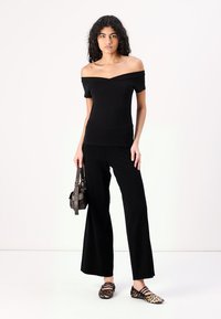 Musta olkaimellinen paita yhdistettynä leveisiin mustiin housuihin. Malli pitää tummasta käsilaukusta kiinni ja päälle on puettu ruskeat ja mustat leopardikuvioiset kengät.