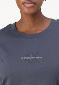 Tmavě šedé bavlněné tričko s kulatým výstřihem. Obsahuje vyšívané logo „CK“ a „Calvin Klein Jeans“ v jemné bílé nitě. Hladká textura.