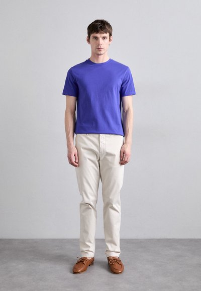 T-shirt en coton violet, col rond, manches courtes. Pantalons beige clair, coupe droite. Chaussures bateau en cuir brun avec lacets.