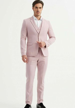 Un costume rose clair avec une coupe ajustée, une veste à un seul bouton, des revers crantés, et un pantalon assorti. Une chemise blanche et des mocassins noirs complètent le look.