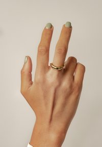 Złoty pierścionek o gładkim, zakrzywionym wzorze na smukłym palcu, z zielonym lakierem do paznokci na dwóch paznokciach i neutralnym tle.